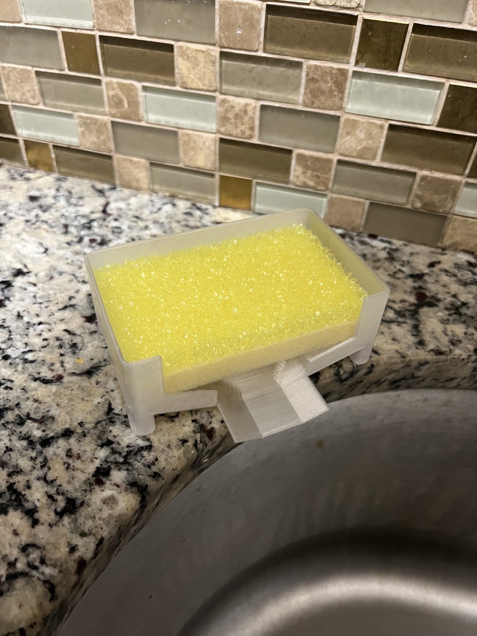 First sponge holder prototype in white with a yellow sponge, on the counter next to the sink