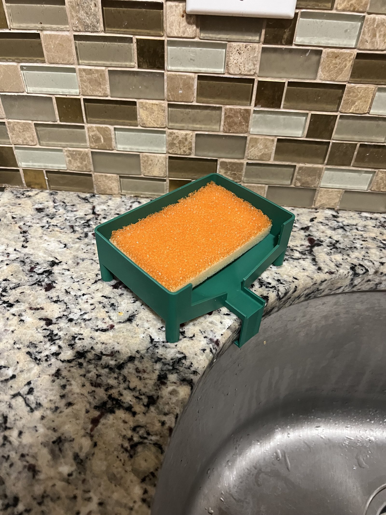 Final green sponge holder with a larger basin, hooked over the counter edge above the sink